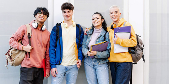 Four international students with folders, backpacks and headphones in front of a white building