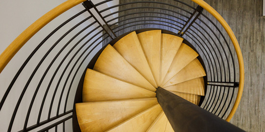 Fast senkrechter Blick von oben auf eine Wendeltreppe mit Holzstufen, Handlauf aus Holz und schwarzem Mittelpfeiler und schwarzen Stahlelementen.