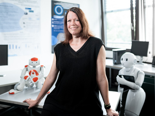 Portrait von Prof. Dr. Nicole Krämer, im Hintergrund stehen zwei Roboter