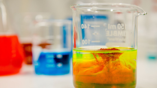 Close-up of a laboratory vessel in which a red and a yellow liquid are mixed, in the background other vessels with colored liquids