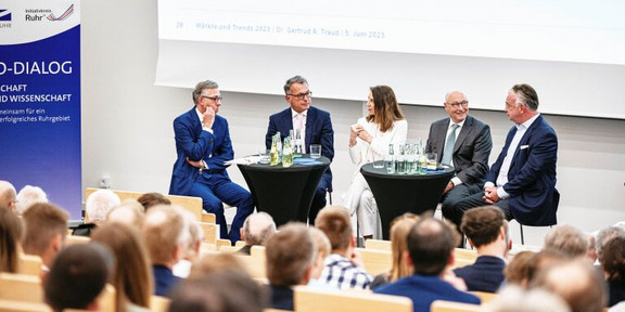 Foto: Eine Gruppe von Menschen spricht auf der Bühne bei einer Podiumsdiskussion. In der Mitte sitzt eine Frau in weißem Anzug und links und rechts von ihr sitzen jeweils zwei Männer in Anzügen.