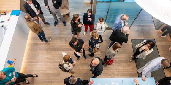 A photo from a bird's eye view. The photo shows a room with bar tables where people are standing together and chatting.