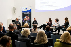 Foto: Aufnahme aus der Perspektive des Publikums bei einer Panel-Diskussion. Eine Gruppe von Personen in Anzügen sitzt auf Stühlen auf einer Bühne und diskutiert miteinander.