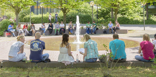 Students sitting on the concrete walls around the ground fountain on Martin-Schmeisser-Platz in summer.