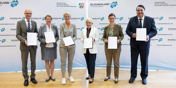 Group photo: Six people in formal dress stand in front of a banner with the inscription Bildungsland NRW and hold certificates in their hands.