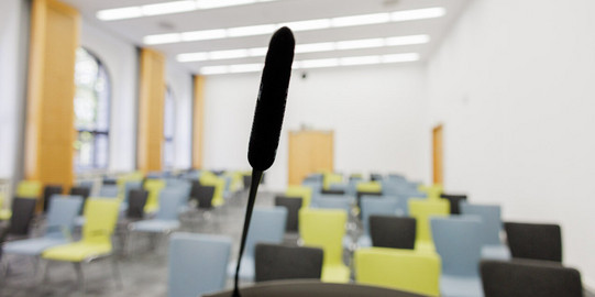 Unscharfer Blick vom Rednerpult in einen Raum mit bunten Stühlen in Reihenbestuhlung im College. Im Vordergrund ist das Mikrofon am Rednerpult im Fokus.