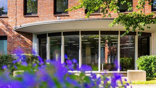 A brick building with a round, glazed entrance area, blurred plants blooming purple in the foreground