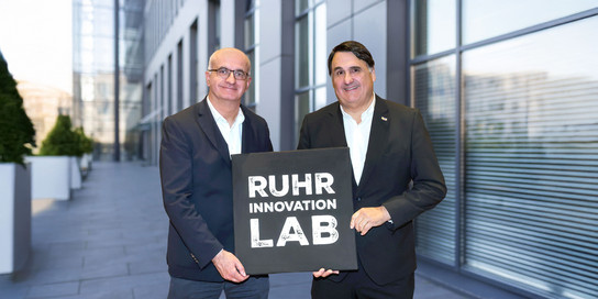 Zwei Rektoren halten ein schwarzes Schild mit der Aufschrift "Ruhr Innovation Lab" hoch.
