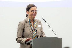 Foto: Eine Frau mit Brille und beigem Anzug steht am Redepult und hält eine Begrüßungsrede.