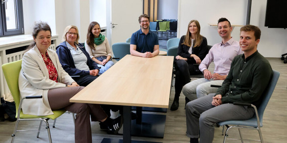 Photo: The seven members of the PostdocLab sit around a table and smile into the camera.